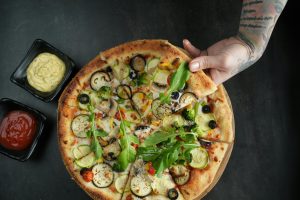 Top view of a vegetarian pizza with fresh toppings and sauces, held by a tattooed hand.