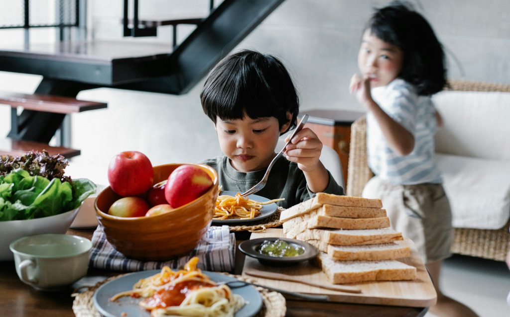 Children eating a homemade lunch with fresh apples, bread, and spaghetti.